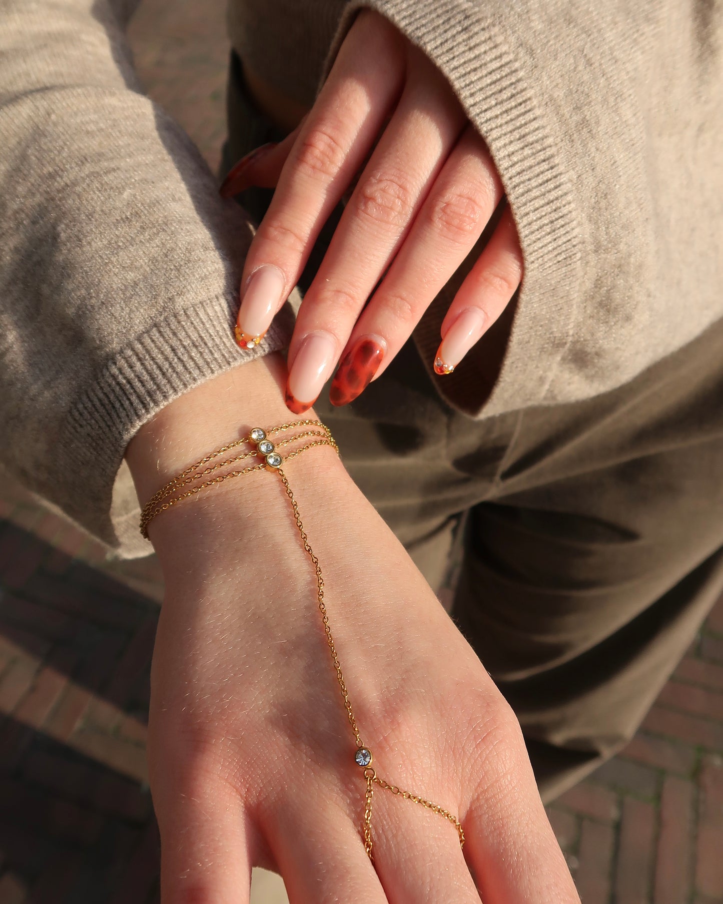 Gouden hand chain met drie fijne armbandbandjes en kleine glinsterende steentjes, verbonden met een ring met één subtiel steentje.