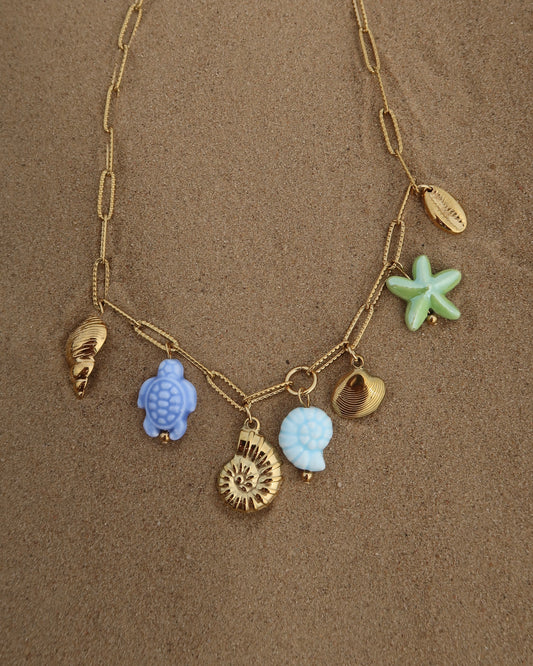 Gouden ketting met zeven bedels, waaronder gouden schelpen, een blauwe schildpad, een blauwe schelp en een groene zeester. De bedels vormen een speelse combinatie van goud met frisse blauw- en groentinten.