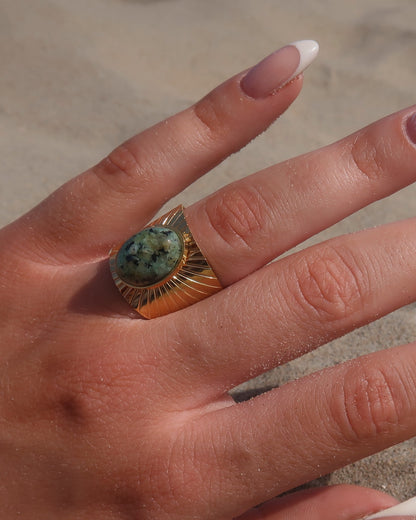 Gouden statement ring van stainless steel met grote ovaalvormige groen gemarmerde steen en subtiel streepjesreliëf in de band.
