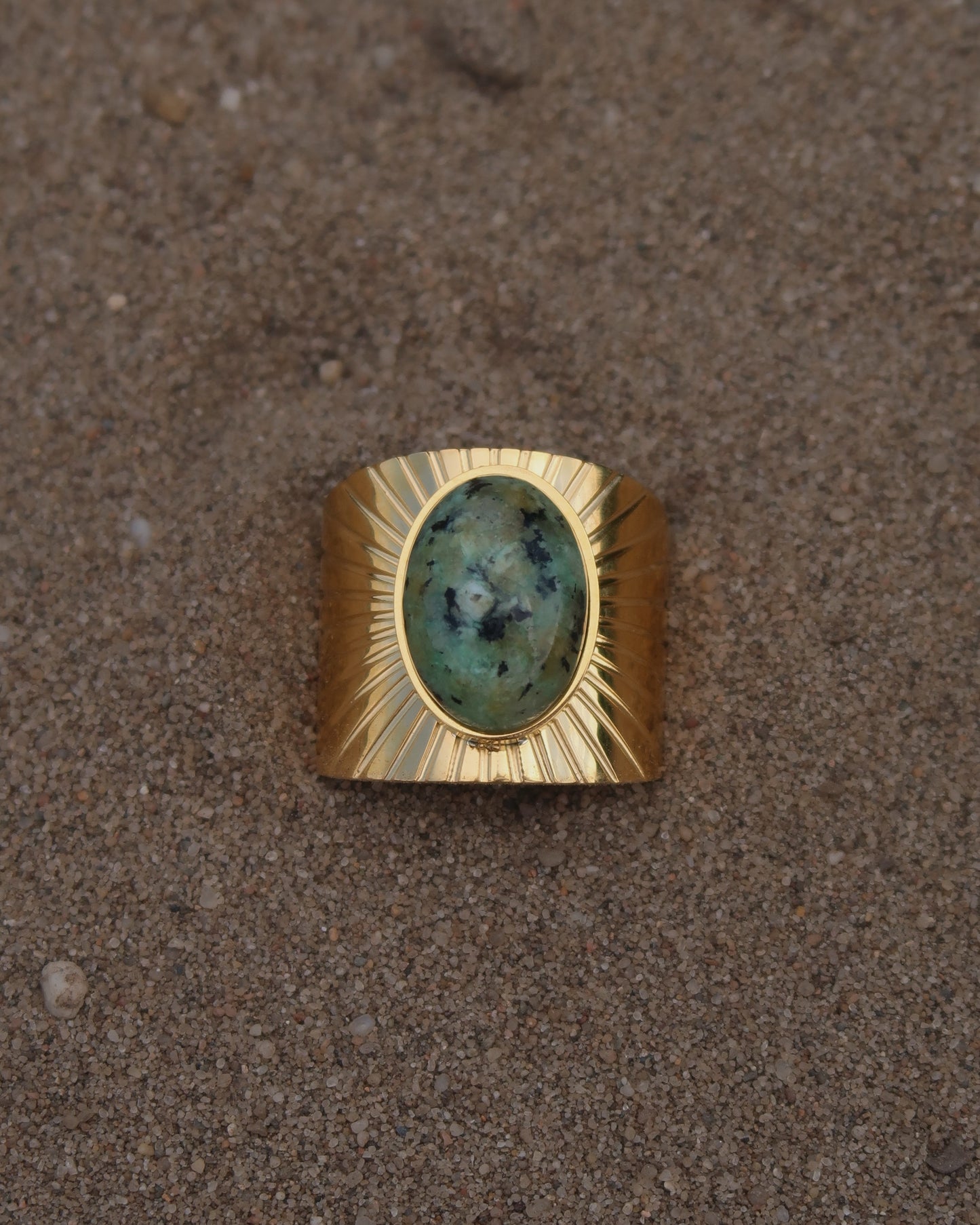 Gouden statement ring van stainless steel met grote ovaalvormige groen gemarmerde steen en subtiel streepjesreliëf in de band.
