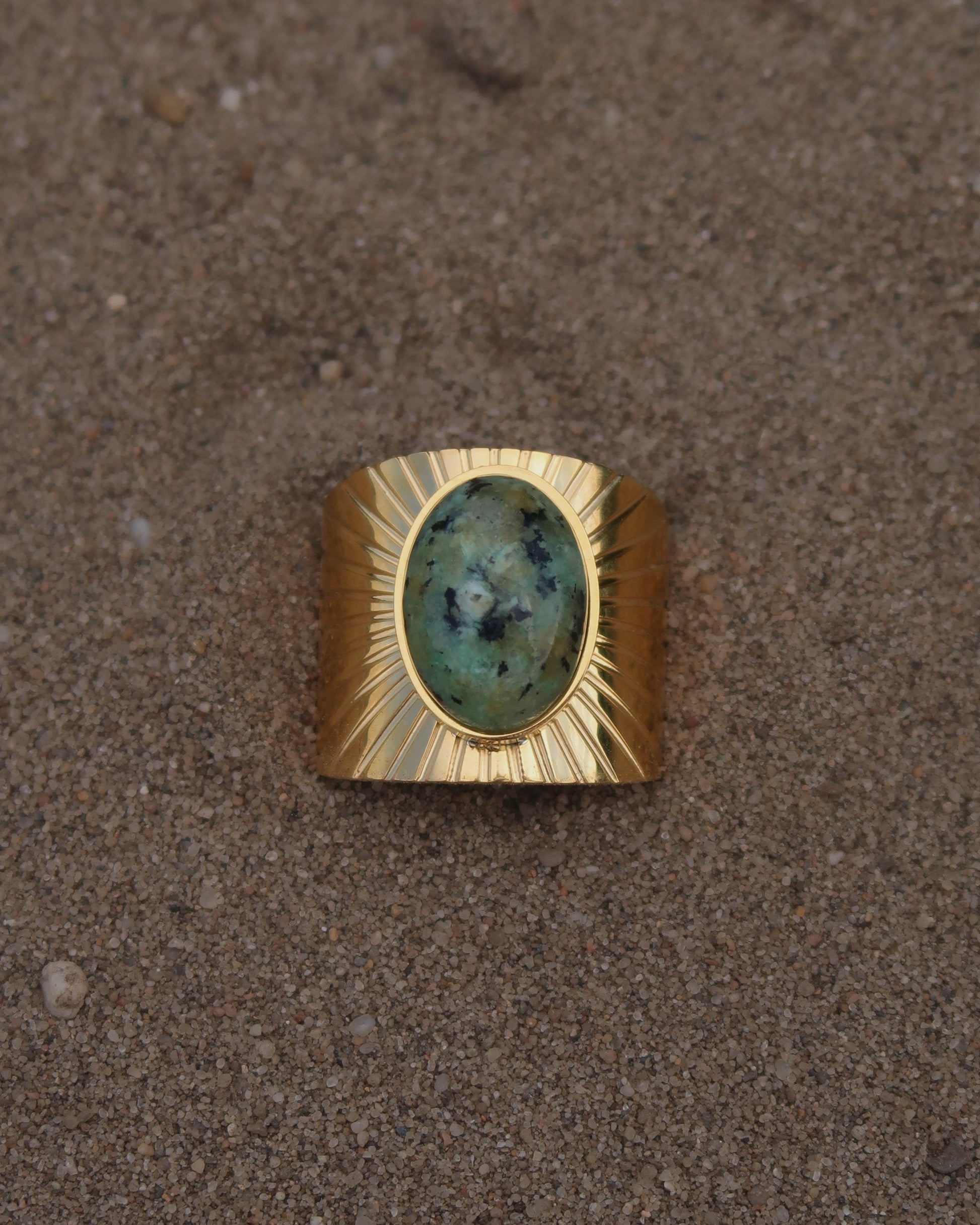 Gouden statement ring van stainless steel met grote ovaalvormige groen gemarmerde steen en subtiel streepjesreliëf in de band.
