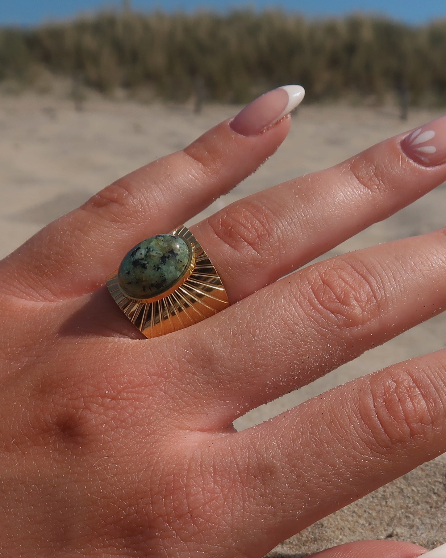 Gouden statement ring van stainless steel met grote ovaalvormige groen gemarmerde steen en subtiel streepjesreliëf in de band.
