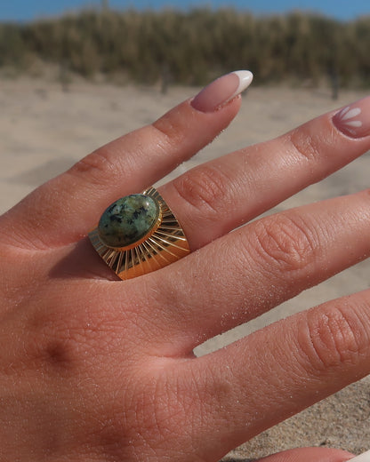 Gouden statement ring van stainless steel met grote ovaalvormige groen gemarmerde steen en subtiel streepjesreliëf in de band.
