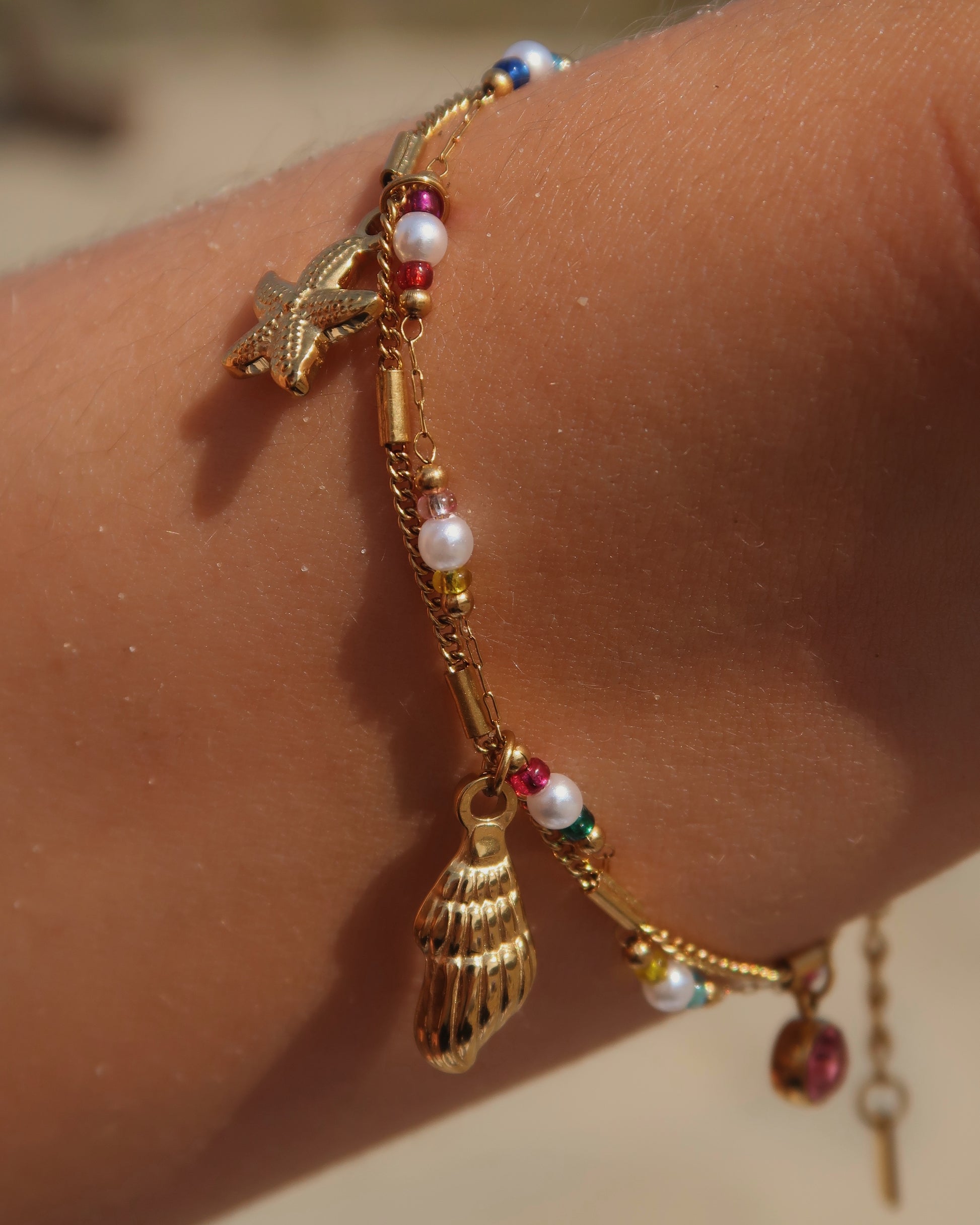 Gouden stainless steel armband met kleurrijke kraaltjes en bedels in de vorm van een schelp, zeester en roze steen – zomerse sieradenstijl.