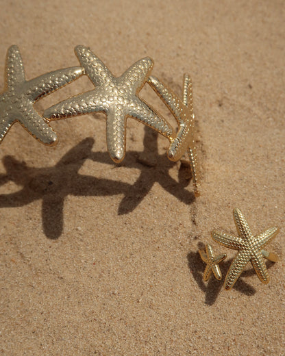 Gouden sieradenset bestaande uit een armband en ring met gedetailleerd zeesterren-design, afgebeeld op zand.