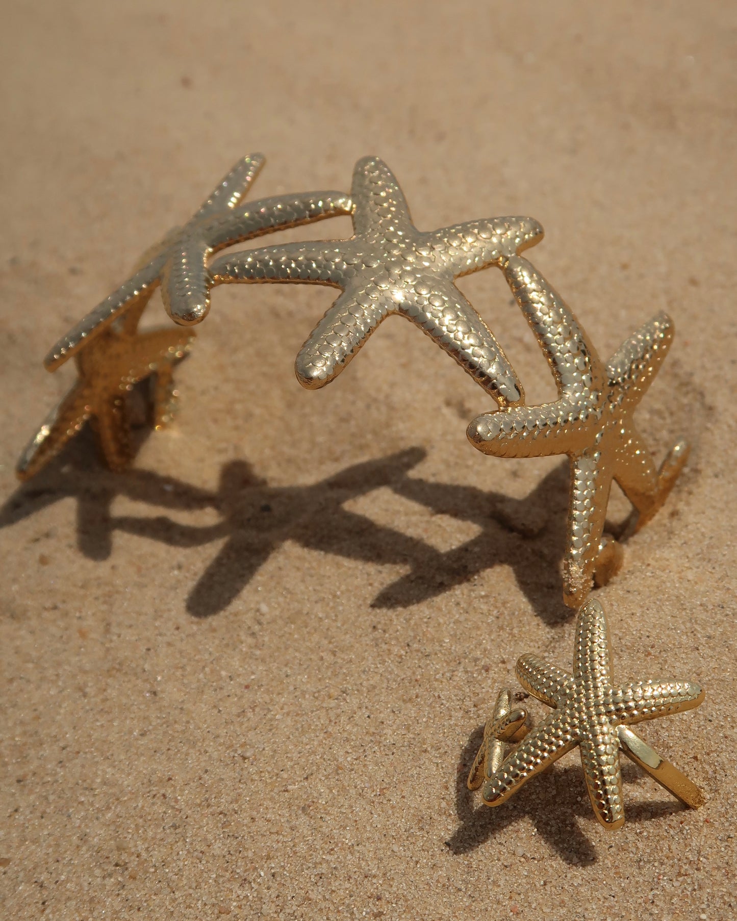 Gouden sieradenset bestaande uit een armband en ring met gedetailleerd zeesterren-design, afgebeeld op zand.