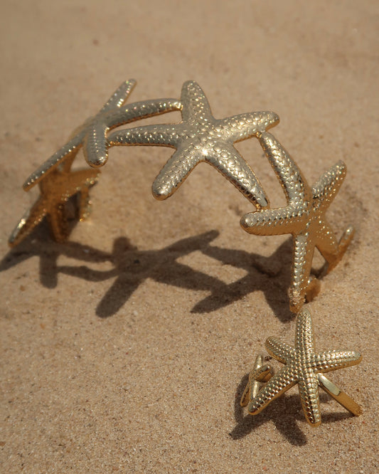 Gouden sieradenset bestaande uit een armband en ring met gedetailleerd zeesterren-design, afgebeeld op zand.