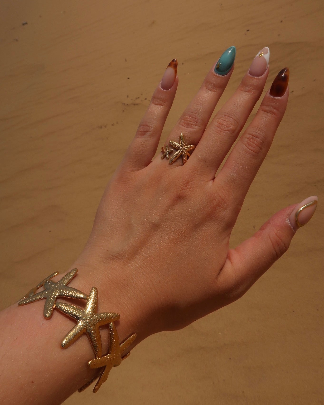Gouden sieradenset bestaande uit een armband en ring met gedetailleerd zeesterren-design, afgebeeld op zand.