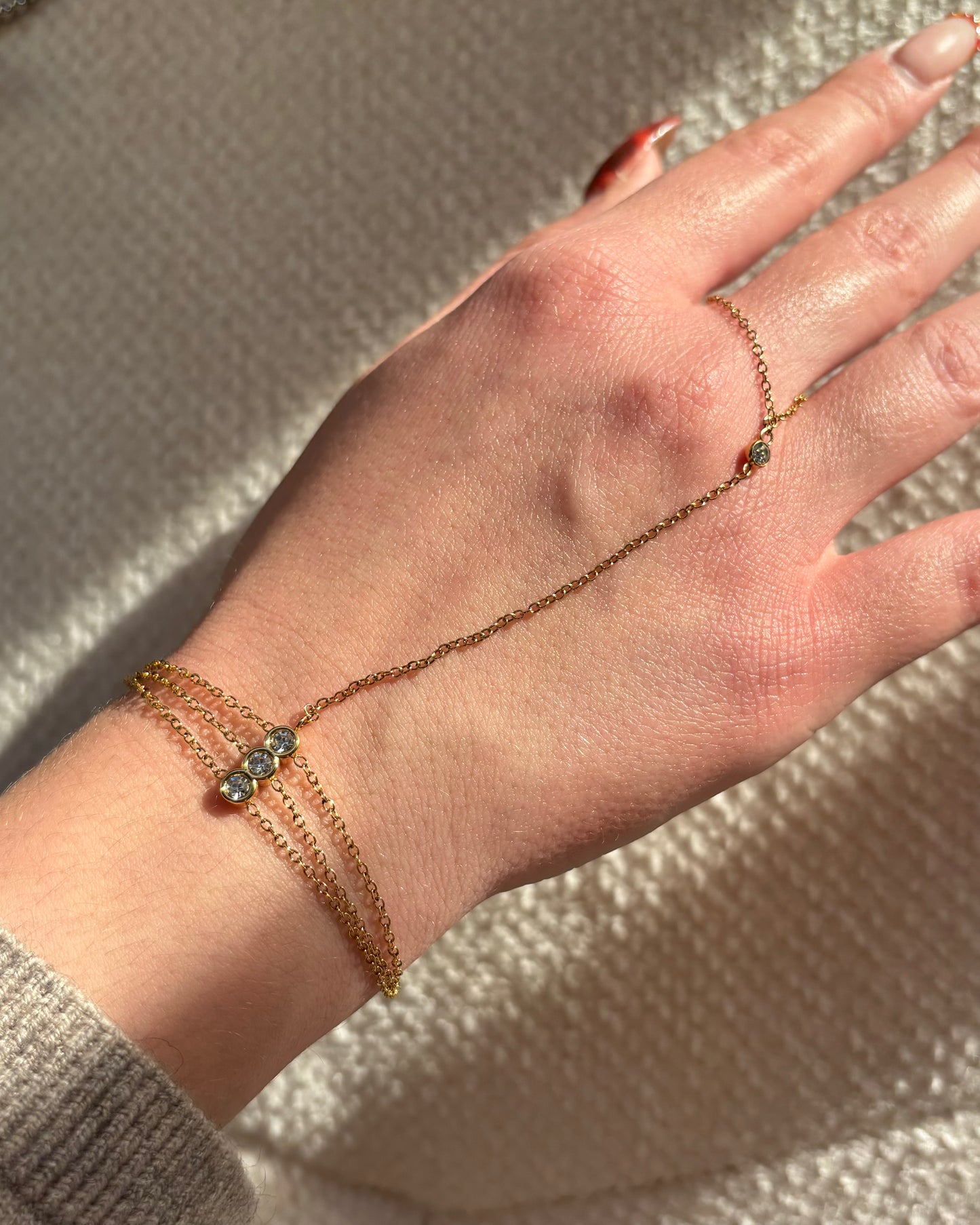 Gouden hand chain met drie fijne armbandbandjes en kleine glinsterende steentjes, verbonden met een ring met één subtiel steentje.