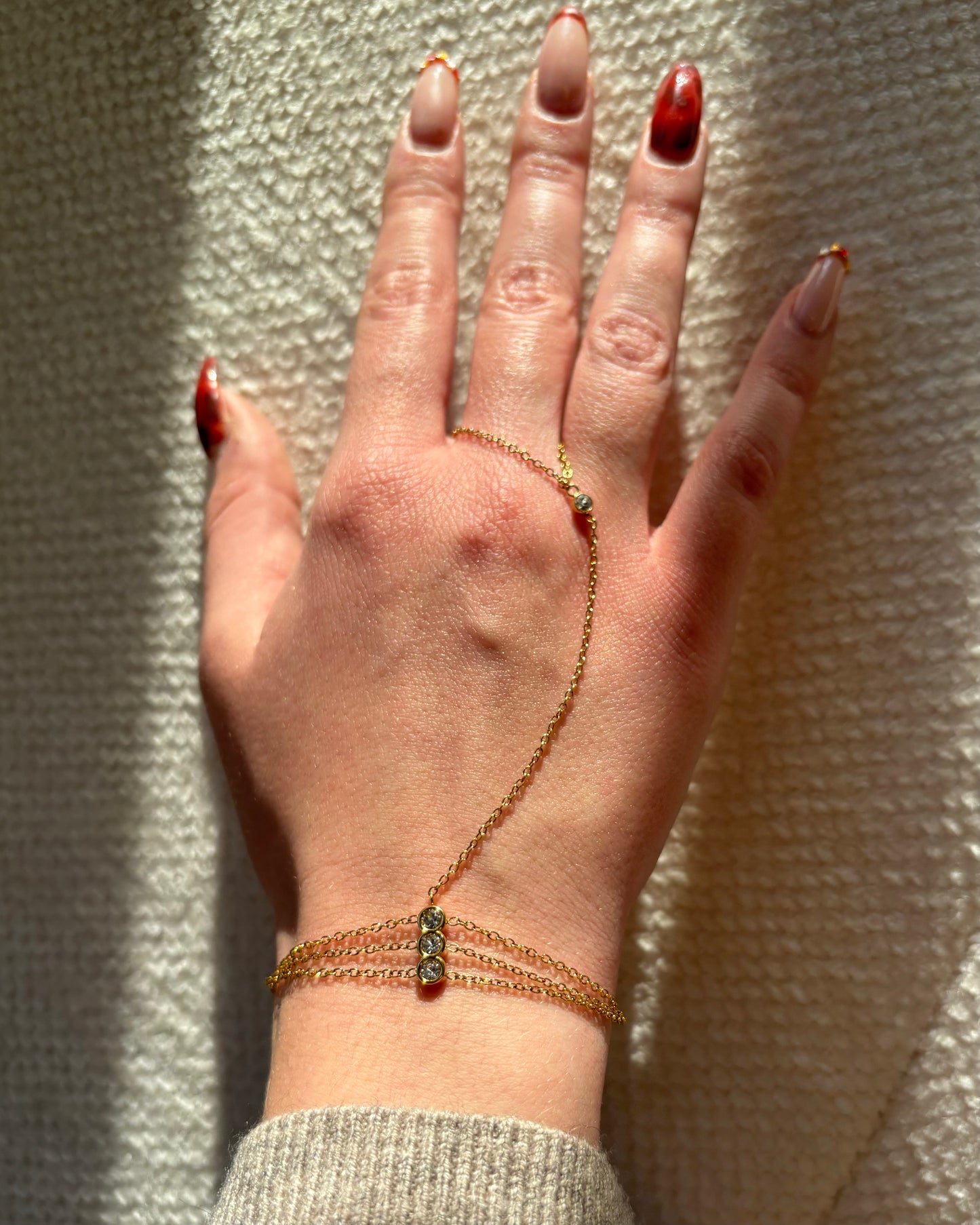 Gouden hand chain met drie fijne armbandbandjes en kleine glinsterende steentjes, verbonden met een ring met één subtiel steentje.