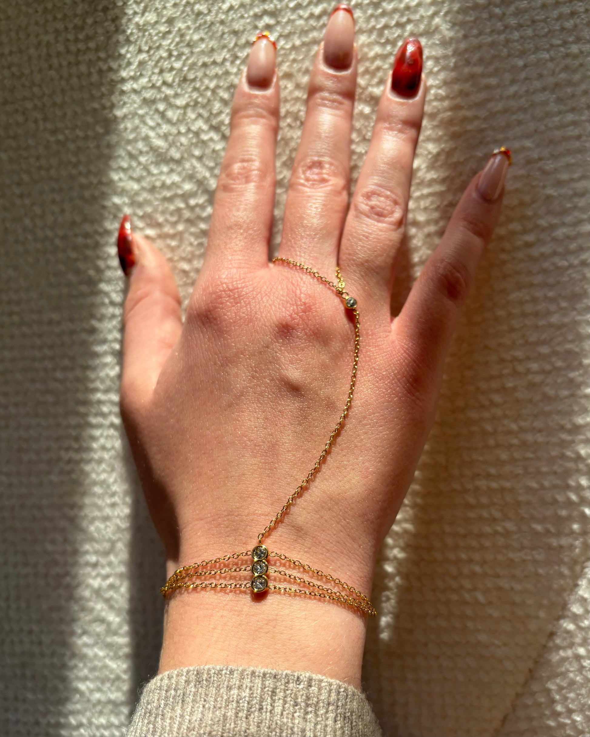 Gouden hand chain met drie fijne armbandbandjes en kleine glinsterende steentjes, verbonden met een ring met één subtiel steentje.