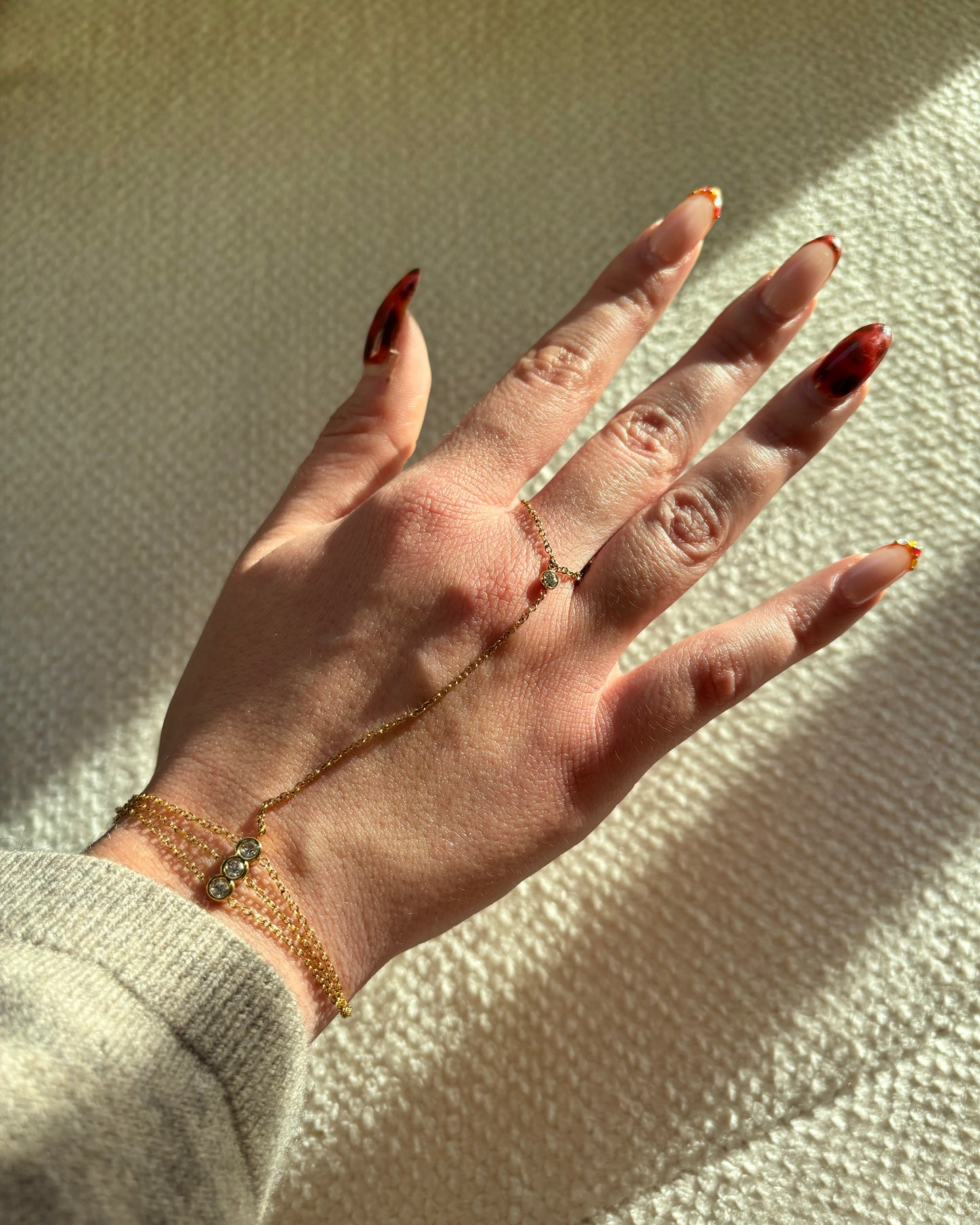 Gouden hand chain met drie fijne armbandbandjes en kleine glinsterende steentjes, verbonden met een ring met één subtiel steentje.