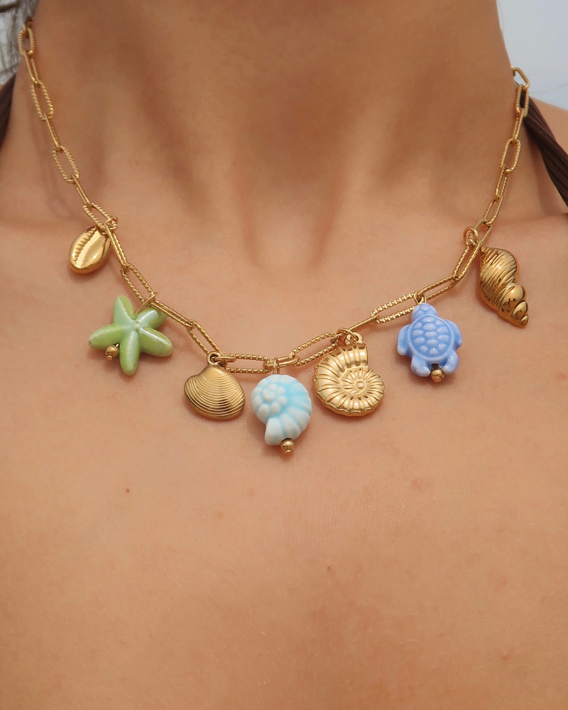 Gouden ketting met zeven bedels, waaronder gouden schelpen, een blauwe schildpad, een blauwe schelp en een groene zeester. De bedels vormen een speelse combinatie van goud met frisse blauw- en groentinten.