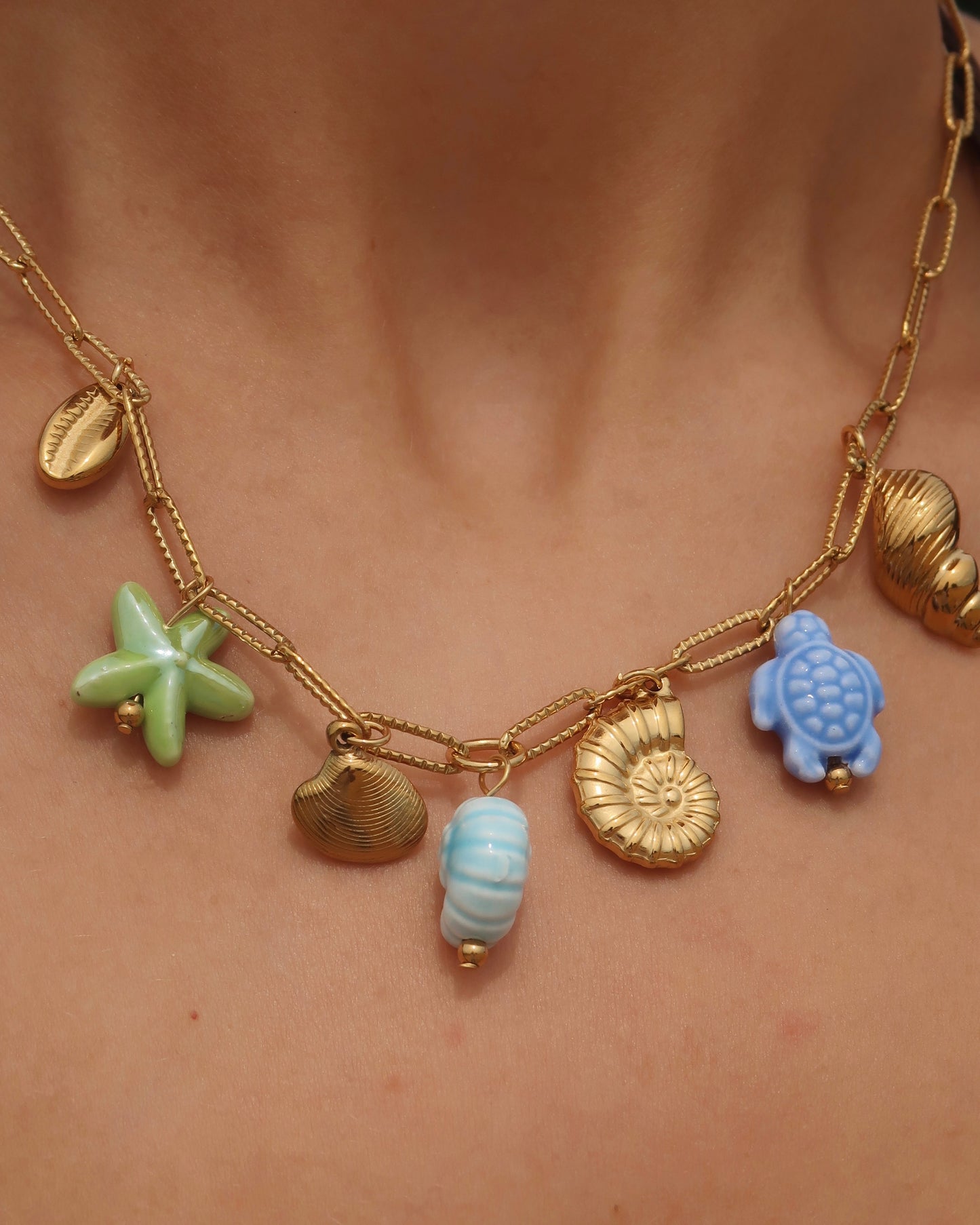 Gouden ketting met zeven bedels, waaronder gouden schelpen, een blauwe schildpad, een blauwe schelp en een groene zeester. De bedels vormen een speelse combinatie van goud met frisse blauw- en groentinten.
