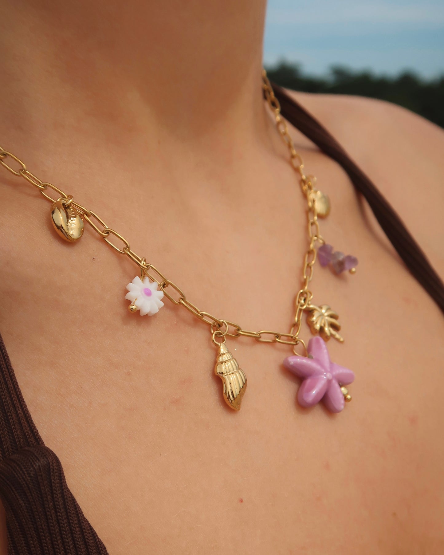 Gouden ketting met zeven bedels, waaronder gouden schelpjes, een paarse zeester, een wit bloemetje met paars hart, paarse steentjes en een gouden palmboom. De ketting heeft een speelse uitstraling met paarse accenten en zomerse details.