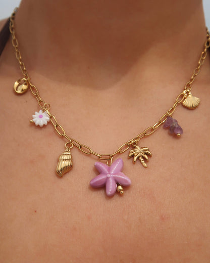 Gouden ketting met zeven bedels, waaronder gouden schelpjes, een paarse zeester, een wit bloemetje met paars hart, paarse steentjes en een gouden palmboom. De ketting heeft een speelse uitstraling met paarse accenten en zomerse details.