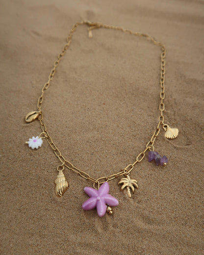 Gouden ketting met zeven bedels, waaronder gouden schelpjes, een paarse zeester, een wit bloemetje met paars hart, paarse steentjes en een gouden palmboom. De ketting heeft een speelse uitstraling met paarse accenten en zomerse details.
