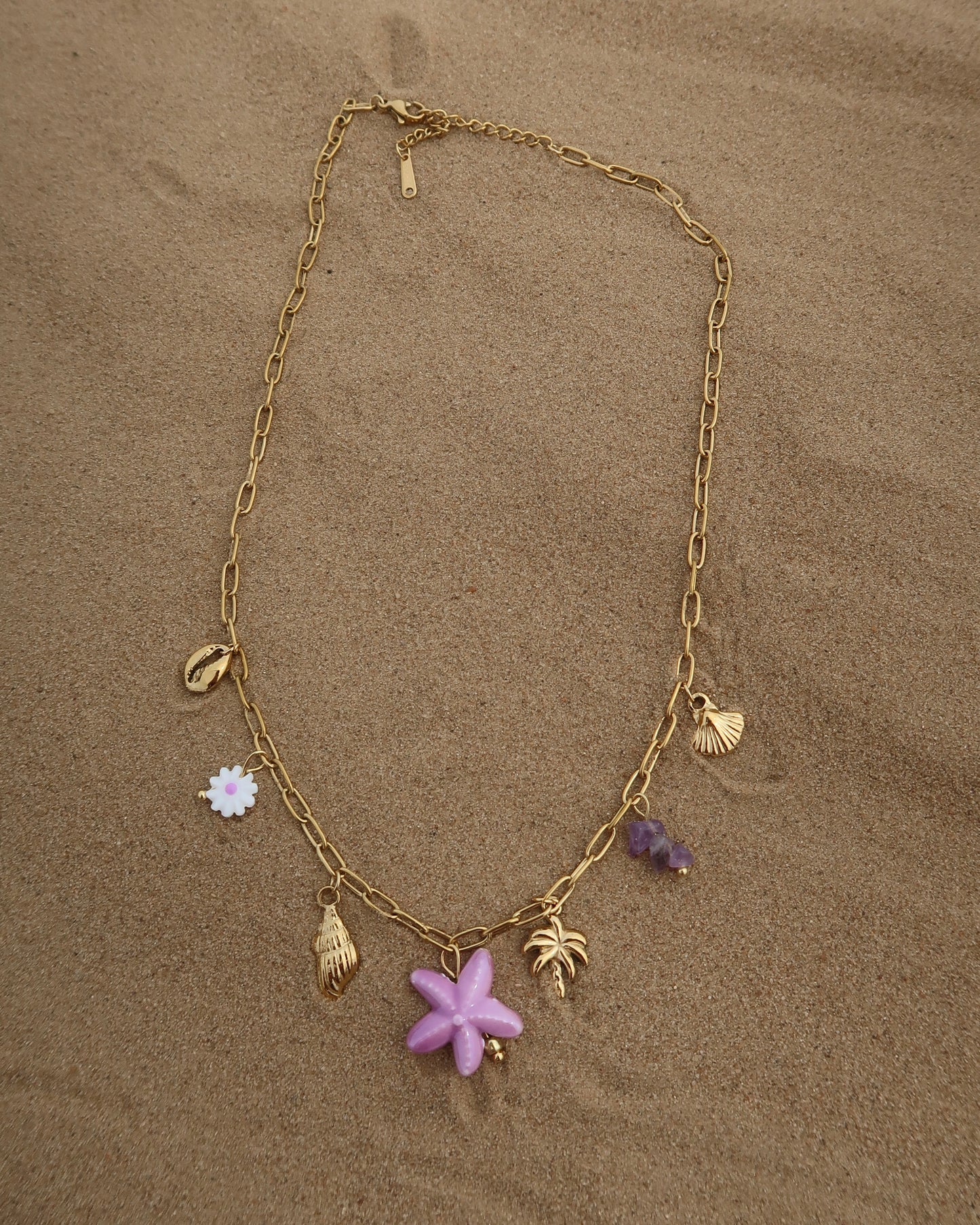 Gouden ketting met zeven bedels, waaronder gouden schelpjes, een paarse zeester, een wit bloemetje met paars hart, paarse steentjes en een gouden palmboom. De ketting heeft een speelse uitstraling met paarse accenten en zomerse details.
