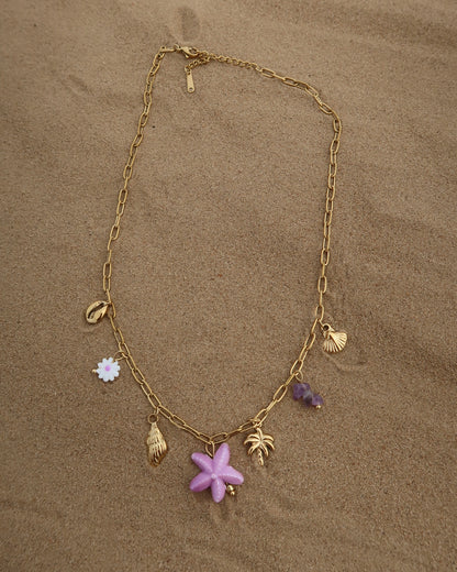 Gouden ketting met zeven bedels, waaronder gouden schelpjes, een paarse zeester, een wit bloemetje met paars hart, paarse steentjes en een gouden palmboom. De ketting heeft een speelse uitstraling met paarse accenten en zomerse details.