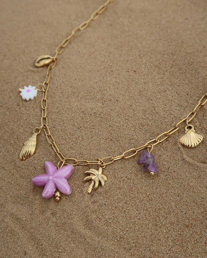 Gouden ketting met zeven bedels, waaronder gouden schelpjes, een paarse zeester, een wit bloemetje met paars hart, paarse steentjes en een gouden palmboom. De ketting heeft een speelse uitstraling met paarse accenten en zomerse details.