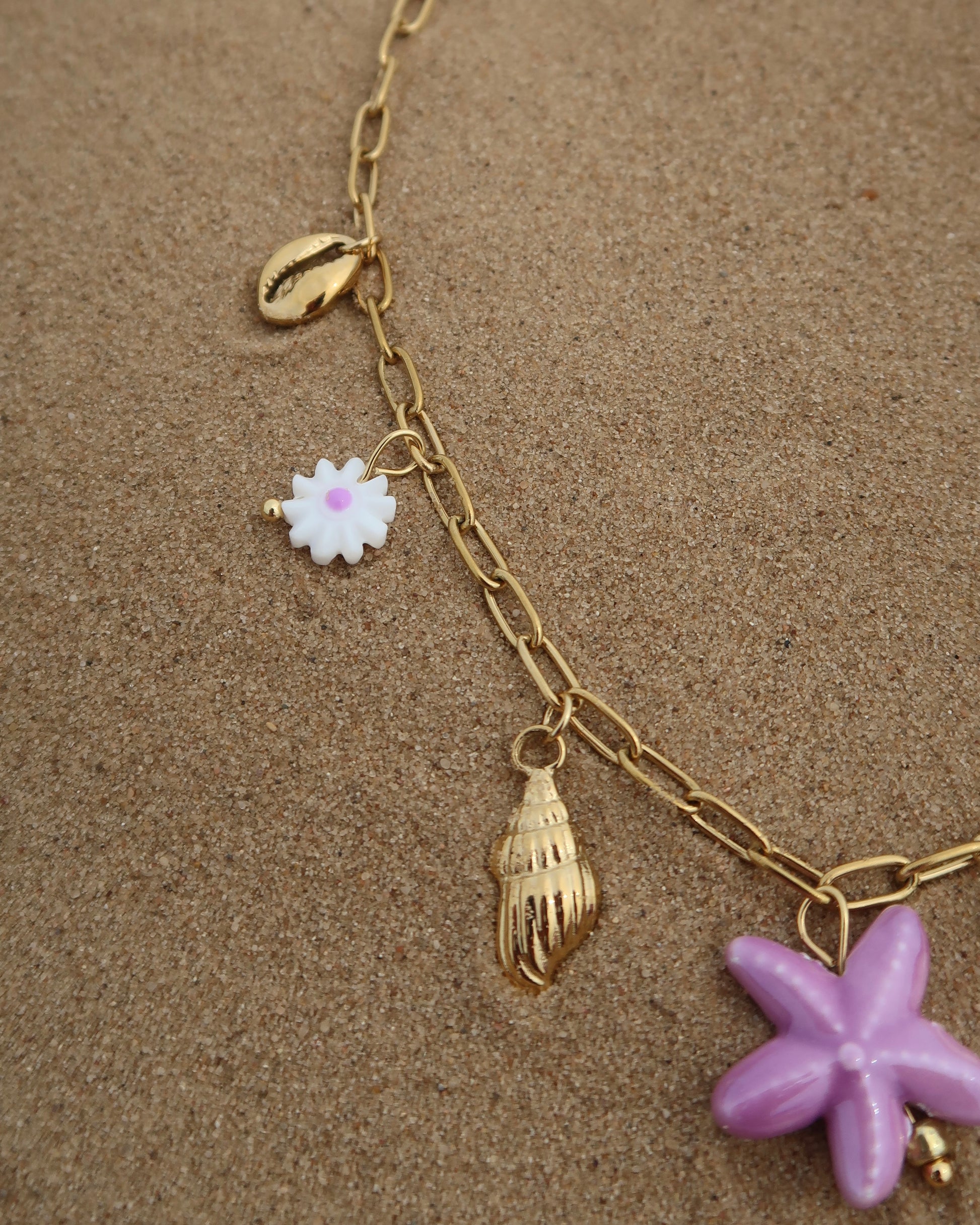 Gouden ketting met zeven bedels, waaronder gouden schelpjes, een paarse zeester, een wit bloemetje met paars hart, paarse steentjes en een gouden palmboom. De ketting heeft een speelse uitstraling met paarse accenten en zomerse details.