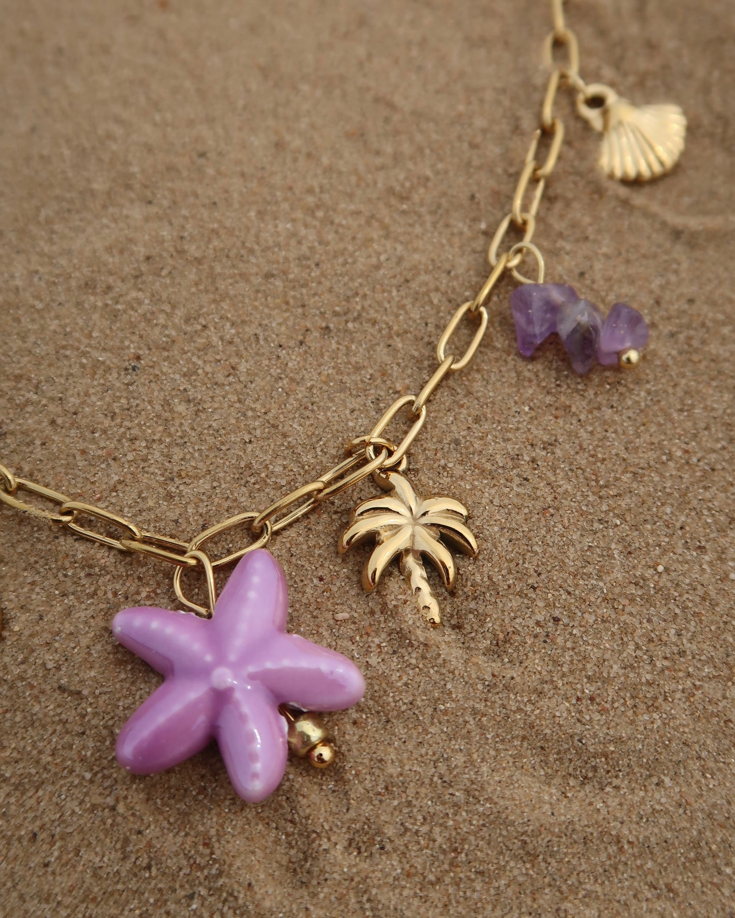 Gouden ketting met zeven bedels, waaronder gouden schelpjes, een paarse zeester, een wit bloemetje met paars hart, paarse steentjes en een gouden palmboom. De ketting heeft een speelse uitstraling met paarse accenten en zomerse details.