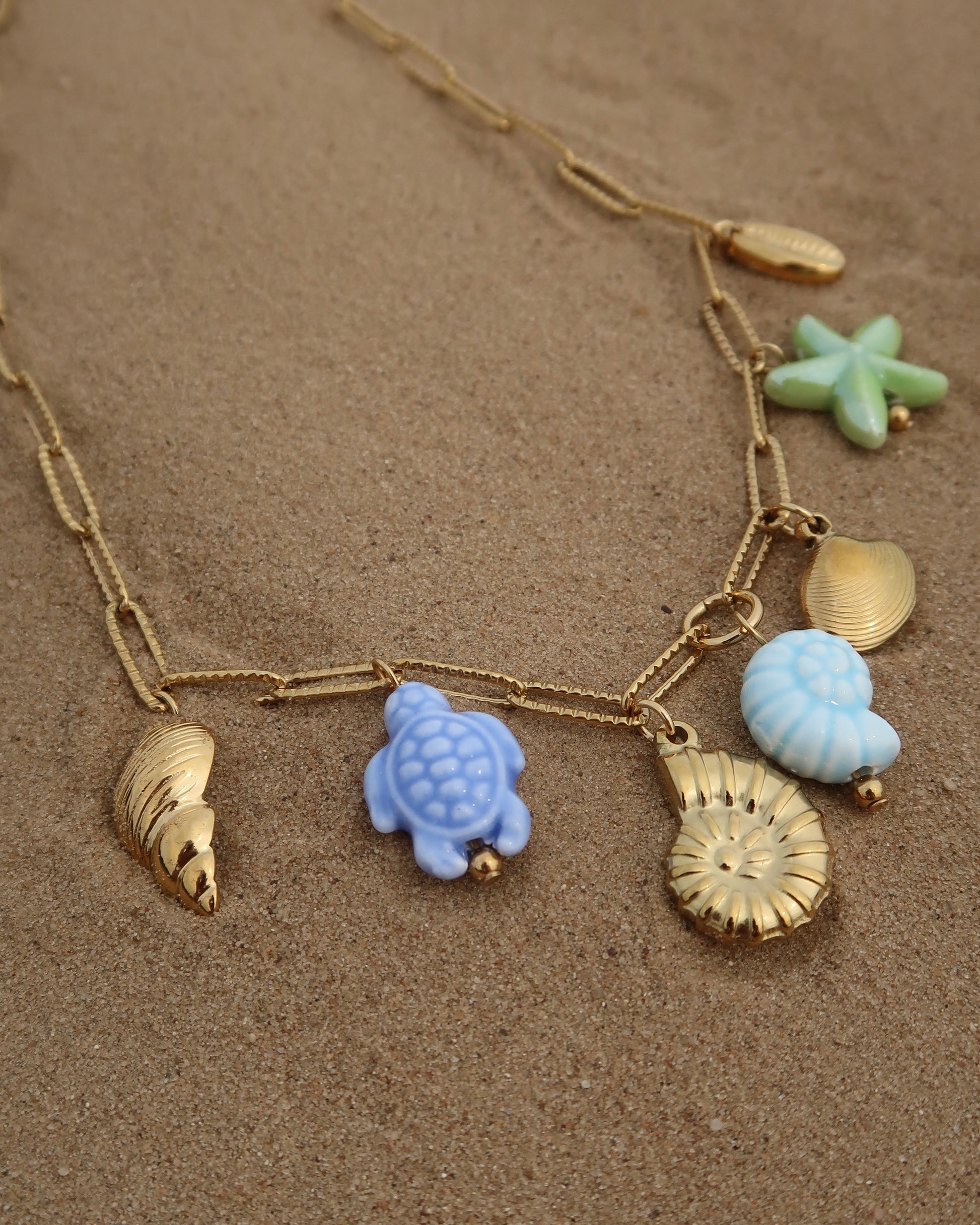 Gouden ketting met zeven bedels, waaronder gouden schelpen, een blauwe schildpad, een blauwe schelp en een groene zeester. De bedels vormen een speelse combinatie van goud met frisse blauw- en groentinten.