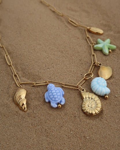 Gouden ketting met zeven bedels, waaronder gouden schelpen, een blauwe schildpad, een blauwe schelp en een groene zeester. De bedels vormen een speelse combinatie van goud met frisse blauw- en groentinten.