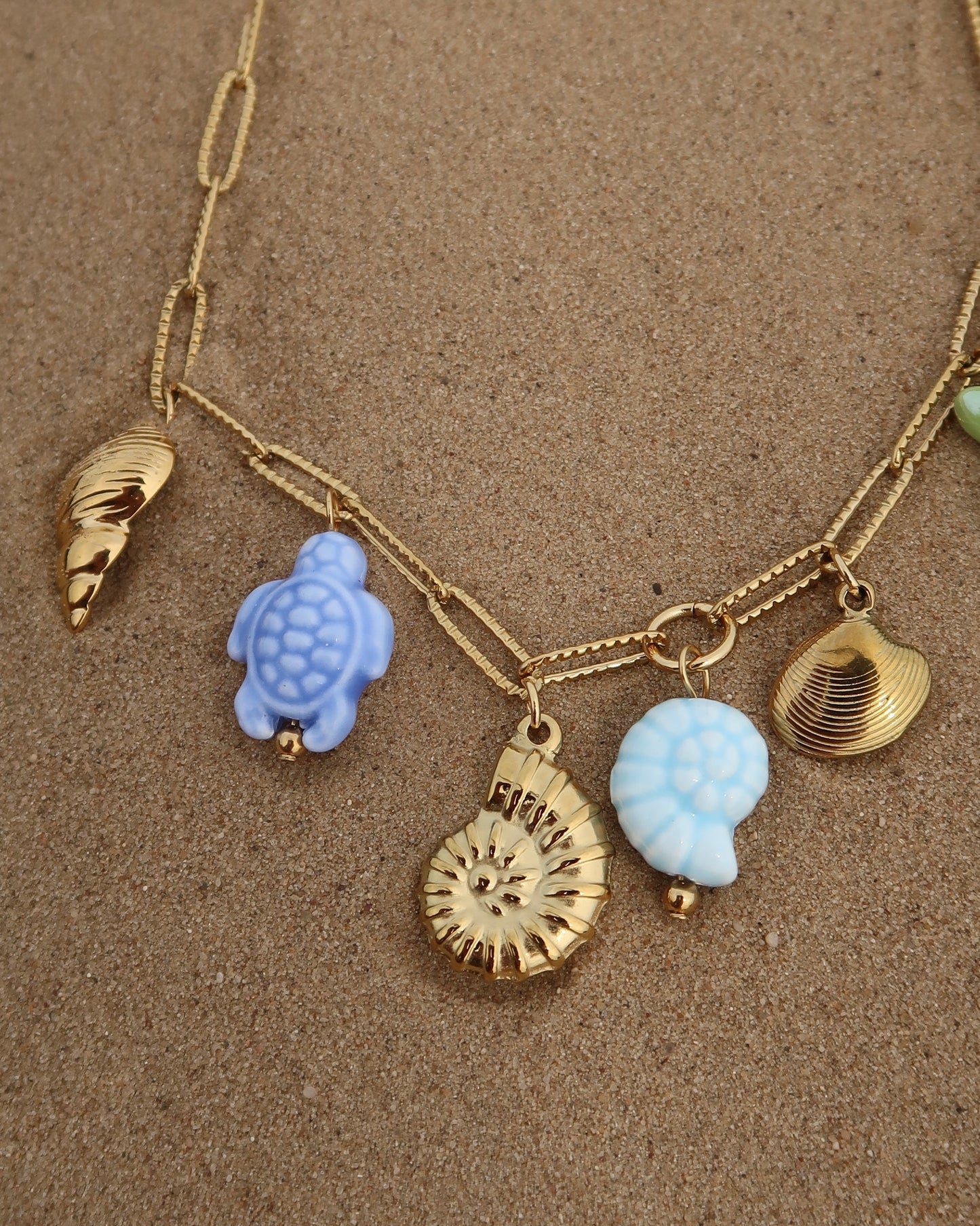 Gouden ketting met zeven bedels, waaronder gouden schelpen, een blauwe schildpad, een blauwe schelp en een groene zeester. De bedels vormen een speelse combinatie van goud met frisse blauw- en groentinten.