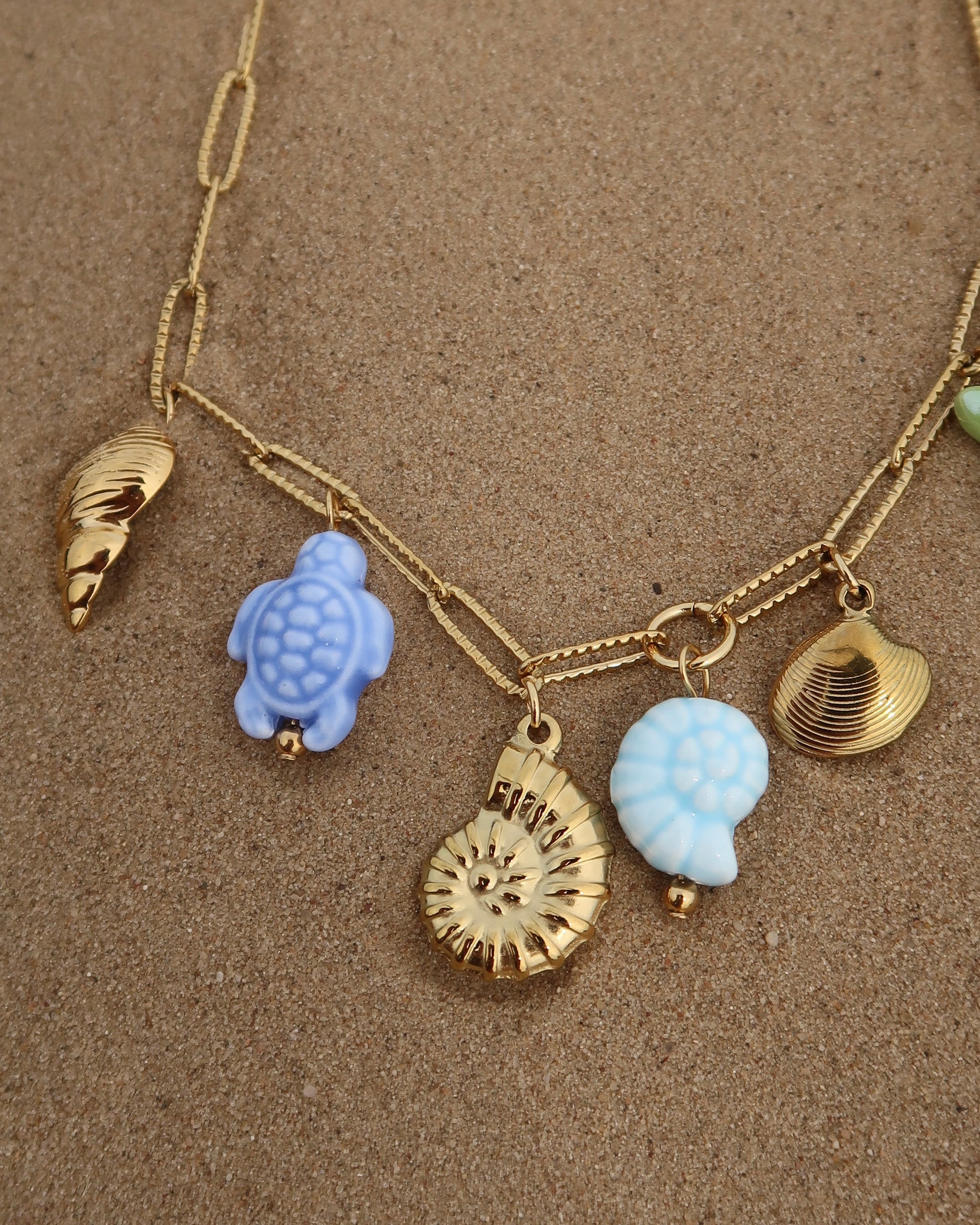 Gouden ketting met zeven bedels, waaronder gouden schelpen, een blauwe schildpad, een blauwe schelp en een groene zeester. De bedels vormen een speelse combinatie van goud met frisse blauw- en groentinten.