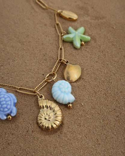 Gouden ketting met zeven bedels, waaronder gouden schelpen, een blauwe schildpad, een blauwe schelp en een groene zeester. De bedels vormen een speelse combinatie van goud met frisse blauw- en groentinten.