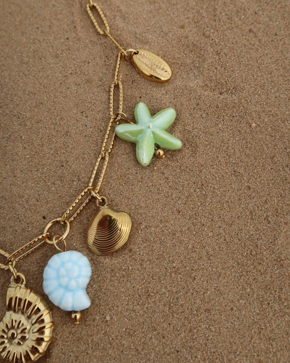 Gouden ketting met zeven bedels, waaronder gouden schelpen, een blauwe schildpad, een blauwe schelp en een groene zeester. De bedels vormen een speelse combinatie van goud met frisse blauw- en groentinten.
